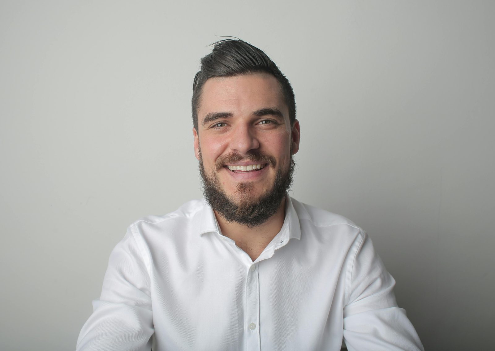 Portrait of a young bearded man smiling, wearing a white shirt indoors against a gray background.